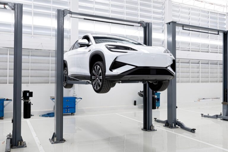 A black two-post car lift raises a white electric vehicle in a modern, bright automotive repair shop.