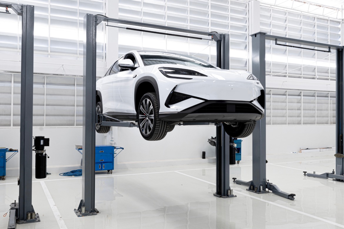 A black two-post car lift raises a white electric vehicle in a modern, bright automotive repair shop.
